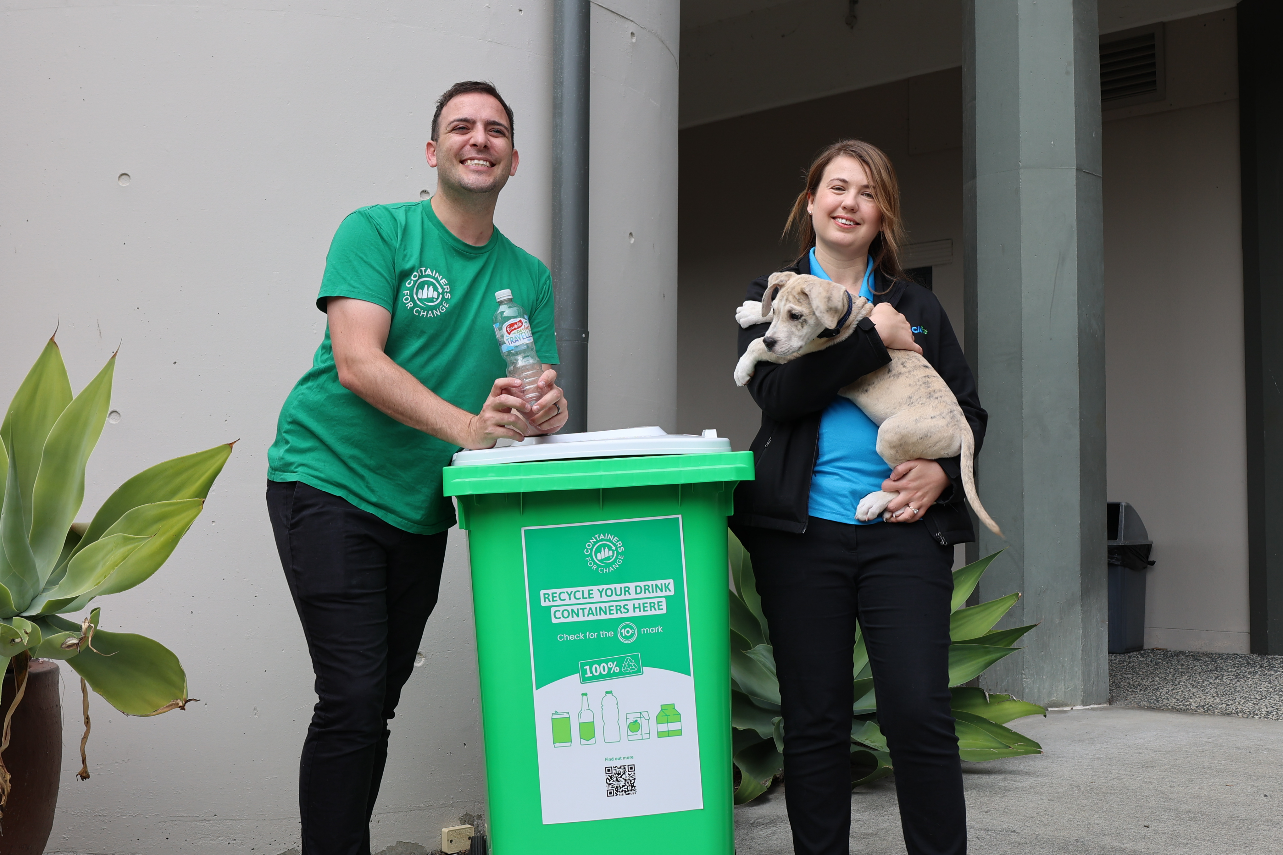 Man, woman and puppy standing in front of green recycling bin