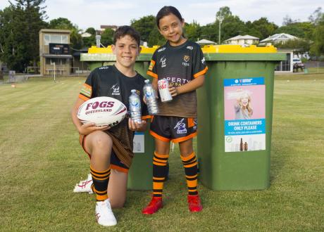 Young boy and girl holding football drink containers, standing beside wheelie bin