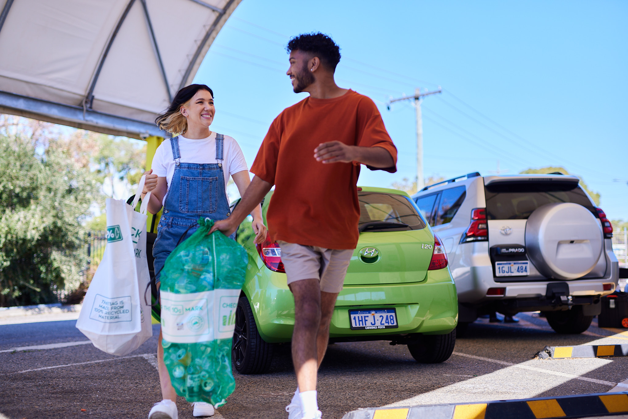 A woman with brown hair, wearing denim overalls and a white shirt is holding a large white bag filled with empty drink containers. A man in a red shirt is beside her, holding a green mesh bag full of empty drink containers.